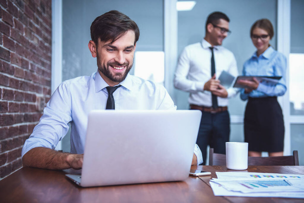 Красивый бизнесмен использует ноутбук и улыбается во время работы в офисе, его коллеги изучают документы на заднем плане
 - Фото, изображение