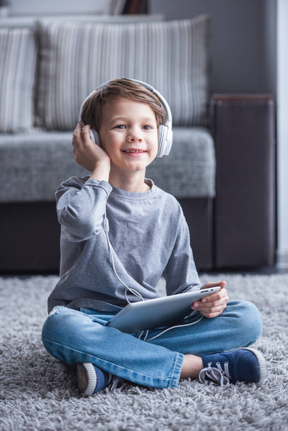 Mały chłopiec w słuchawkach jest słuchanie muzyki przy użyciu cyfrowego tabletu i uśmiechając się podczas gry w domu - Zdjęcie, obraz