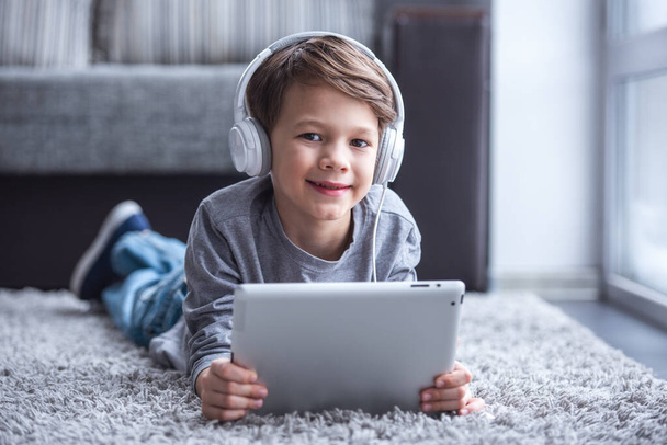 Μικρό αγόρι ακουστικά είναι χρησιμοποιώντας μια ψηφιακή δισκίο, βλέπουν φωτογραφική μηχανή και χαμογελώντας ενώ ξαπλωμένο στο πάτωμα στο σπίτι - Φωτογραφία, εικόνα