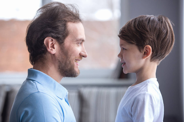 Vista lateral del apuesto padre y su lindo hijo mirándose y sonriendo mientras pasan tiempo juntos en casa
 - Foto, imagen