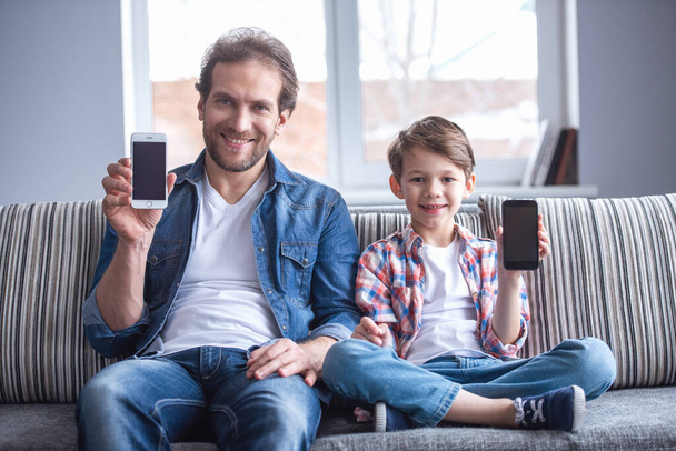 Отец и сын показывают смартфоны, смотрят в камеру и улыбаются, сидя дома на диване.
 - Фото, изображение