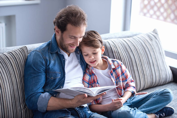 Πατέρας και γιος διαβάζουν ένα βιβλίο και χαμογελούν ενώ περνούν χρόνο μαζί στο σπίτι. - Φωτογραφία, εικόνα