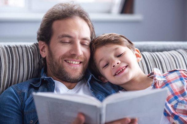 Отец и сын читают книгу и улыбаются, проводя время вместе дома - Фото, изображение