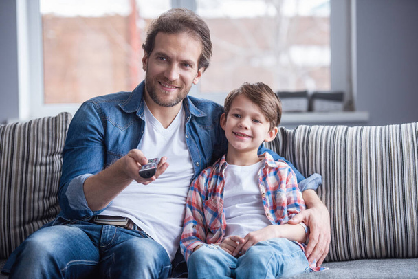 Отец и сын смотрят телевизор и улыбаются, проводя время вместе дома
 - Фото, изображение