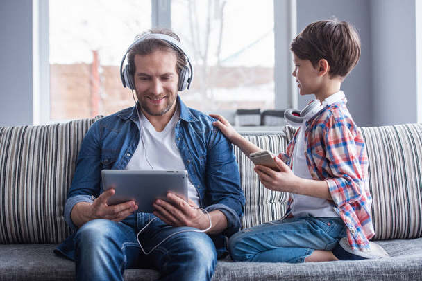 Padre e hijo en auriculares están escuchando música usando aparatos y sonriendo mientras pasan tiempo juntos en casa
 - Foto, Imagen