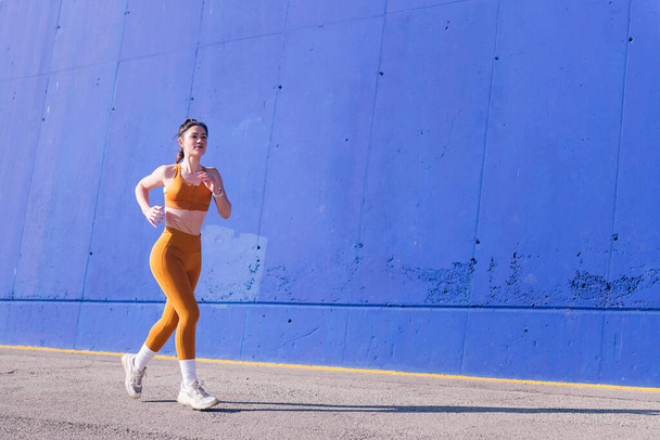 caucasian woman with orange sportswear running in an urban track, concept of sport and active lifestyle, copy space for text - 写真・画像