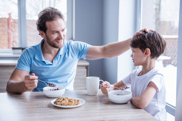 Pai e filho estão conversando e sorrindo enquanto tomam um café da manhã na cozinha
 - Foto, Imagem