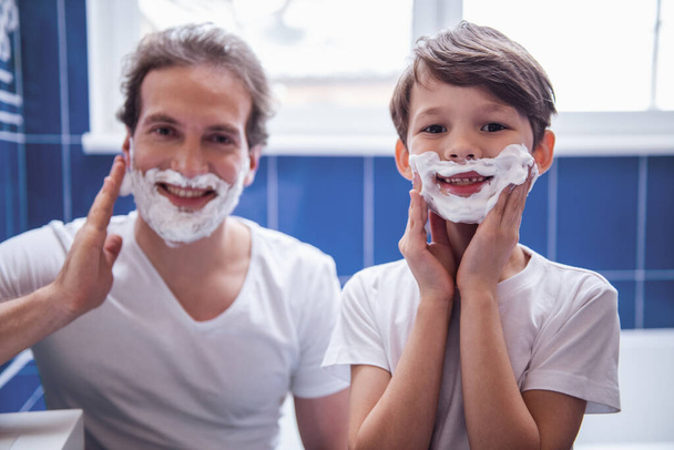 Isä ja poika levittävät partavaahtoa kasvoilleen ja hymyilevät ajaessaan partaa kylpyhuoneessa.
 - Valokuva, kuva