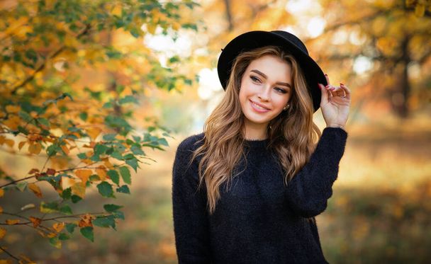 Primer plano retrato de una hermosa chica en vestido oscuro y sombrero negro de pie cerca de coloridas hojas de otoño. Obra de arte de mujer romántica . - Foto, imagen