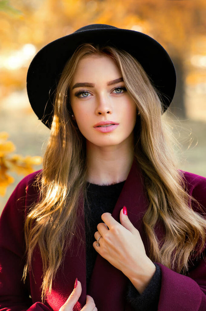 カラフルな秋の葉の近くに立って暗いドレスと黒の帽子の美しい女の子の肖像画を閉じます。ロマンチックな女性のアート作品.カメラを見てかなり優しさモデル. - 写真・画像