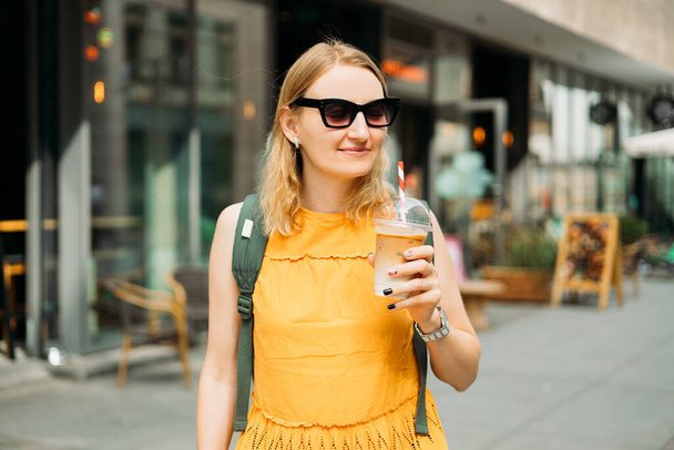 Schöne modische junge Frau mit Sonnenbrille, die auf der Straße der Stadt steht. Urban Lifestylekonzept. Attraktive Frau trinkt einen Eiskaffee in Plastikbecher. - Foto, Bild