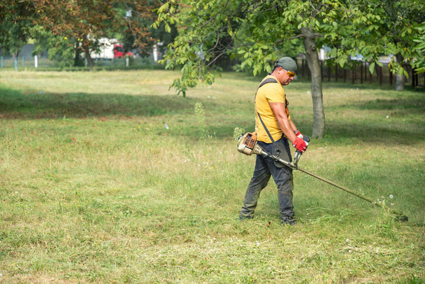 Muž zahradník seká trávu s elektrickým nebo benzínovým sekačem trávníku v parku. Zahradní nářadí a vybavení. Muž seká trávu benzínovou kosou. Copyspace. - Fotografie, Obrázek