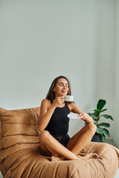 Kapalı gözlü, keyifli genç bir kadın bir fincan kahvenin tadını çıkarıyor, rahat fasulye koltukta oturuyor. - Fotoğraf, Görsel