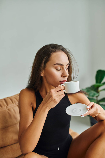Kumral saçlı güzel genç bir kadın sabah kahvesinin tadını çıkarıyor, fasulye sırığında oturuyor. - Fotoğraf, Görsel