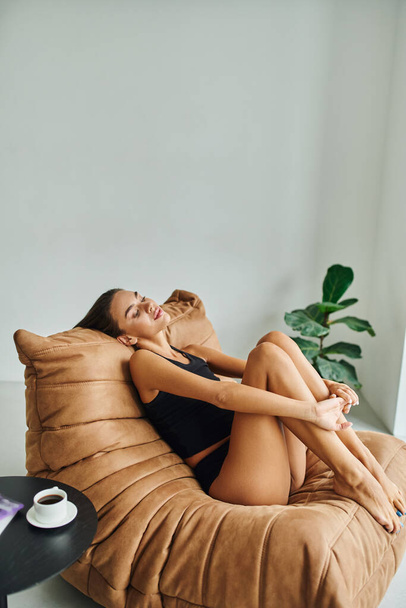 beautiful young woman resting on bean bag chair near coffee table with cup of black coffee - Фото, изображение