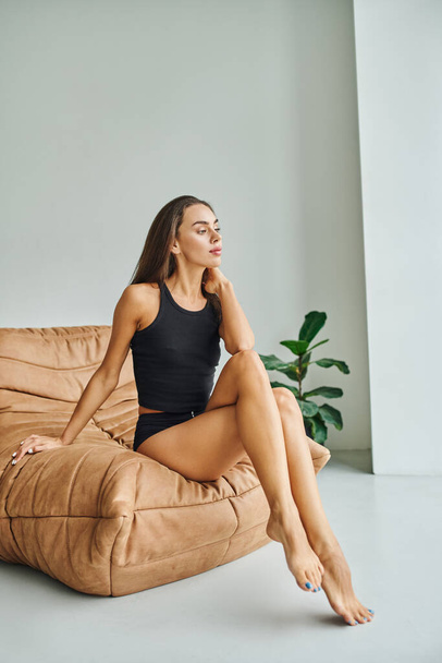 mujer soñadora y descalza sentada en la silla de la bolsa de frijol y la planta verde en el apartamento moderno, mirar hacia otro lado - Foto, Imagen
