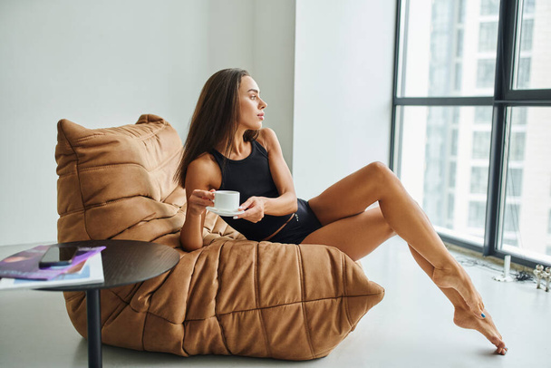barefoot young woman with brunette hair sitting on bean bag chair and holding cup of coffee - Photo, Image