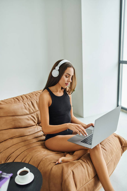 freelance pieds nus dans les écouteurs sans fil en utilisant un ordinateur portable et assis sur une chaise de sac de haricot, jolie femme - Photo, image
