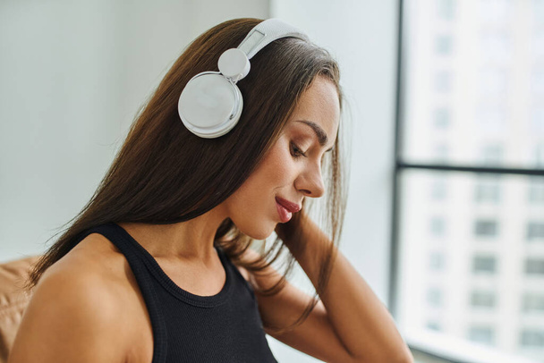 jolie femme brune aux cheveux longs écoutant de la musique dans des écouteurs sans fil à la maison - Photo, image
