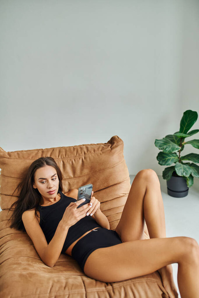 piękna kobieta w koszulce zbiornika i majtki siedzi na krześle worek fasoli i za pomocą smartfona - Zdjęcie, obraz