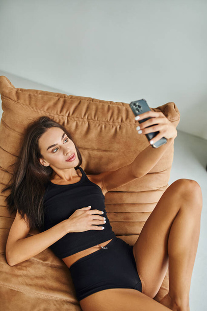hermosa mujer en camiseta sin mangas y bragas descansando en la silla de la bolsa de frijoles y tomando selfie en el teléfono inteligente - Foto, imagen