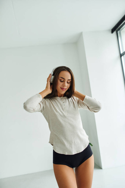 zadowolona brunetka kobieta w długim rękawie i majtki spaceru podczas słuchania muzyki w słuchawkach - Zdjęcie, obraz