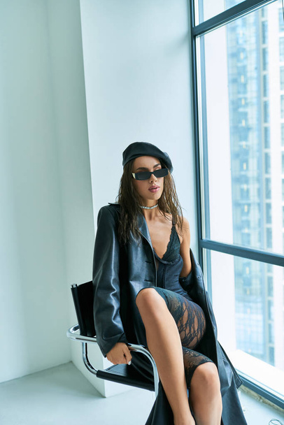 modèle séduisant en lunettes de soleil, béret, sous-vêtements en dentelle et manteau en cuir assis sur la chaise près de la fenêtre - Photo, image