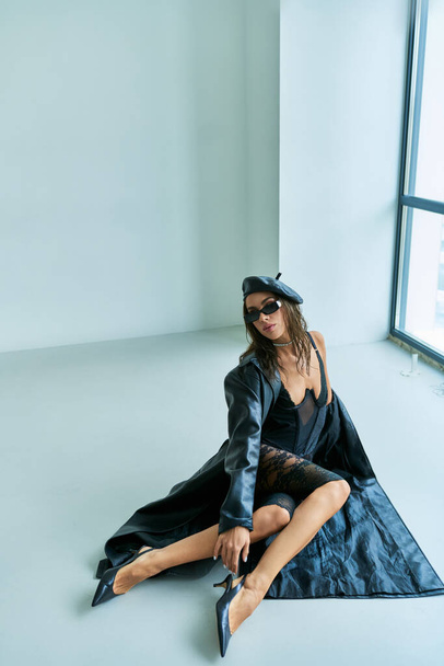 sexy outfit, seductive woman in beret, sunglasses, lace underwear and leather coat sitting on floor - Фото, изображение