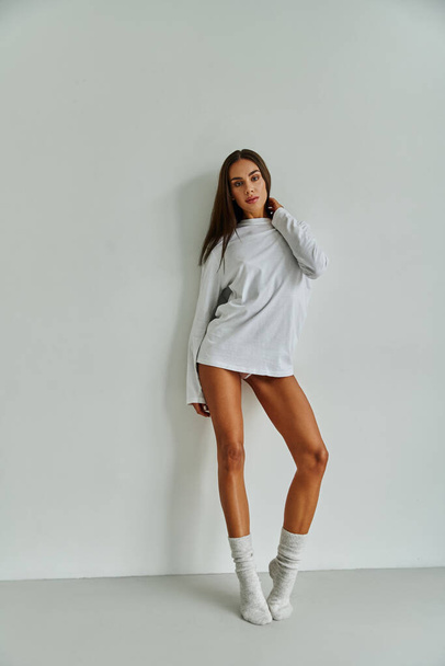 full length of brunette woman with long hair standing in long sleeve and socks near wall at home - Foto, Bild