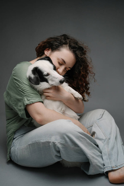 anxious dog hugs.  Beautiful woman with curly hair calming hugging cute sad dog sitting on the floor with dark grey background. Pet mental support. Blue jeans barefoot .  - 写真・画像