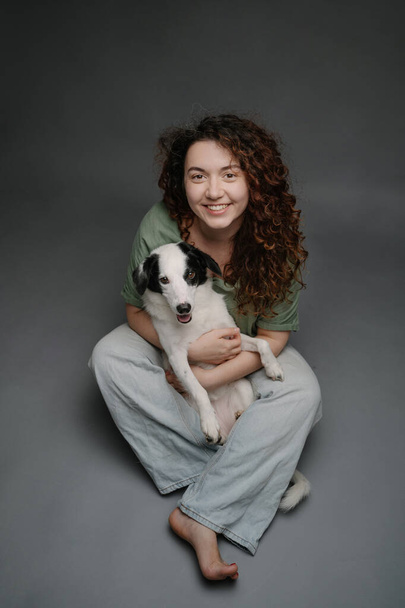 Happy woman hugging her border collie dog happy laughing and looking at the camera. Adorable friends portrait. Grey background studio shot. Sitting on floor in blue jeans barefoot - Photo, image