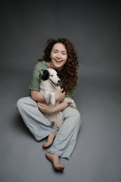 Adorable friends portrait. Happy woman hugging her border collie dog happy laughing and looking at the camera. Grey background studio shot. Sitting on floor in blue jeans barefoot - Photo, image