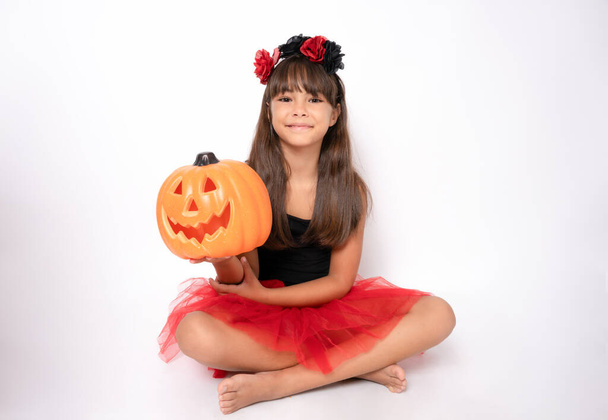 Cute little girl lying on floor in halloween costume holding a pumpkin isolated over white background. - Фото, изображение