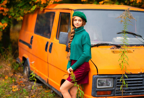 Portret van jong mooi blond meisje met blauwe ogen en gevlochten haar.Aantrekkelijk model in groen gebreide trui en baret staande in de buurt van oude retro oranje bus met herfst achtergrond. Kleurrijke kunst. - Foto, afbeelding
