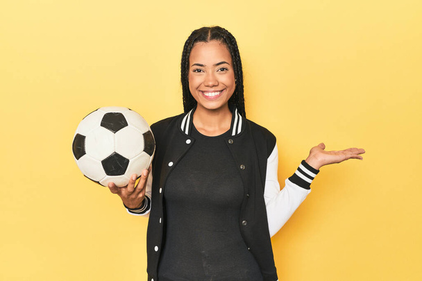 Indonéská školačka s fotbalovým míčem na žluté ukazující kopírovací prostor na dlani a držící další ruku v pase. - Fotografie, Obrázek