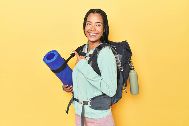 Indonesian woman ready for mountain trip on yellow smiling and pointing aside, showing something at blank space. - Photo, Image