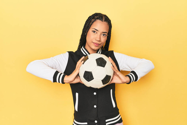 黄色いスタジオの背景にメダルとサッカーボールを持つ若いインドネシアの女性 - 写真・画像
