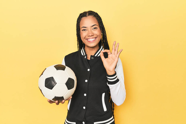 Индонезийская школьница с футбольным мячом на желтый веселый и уверенно показывая ОК жест. - Фото, изображение