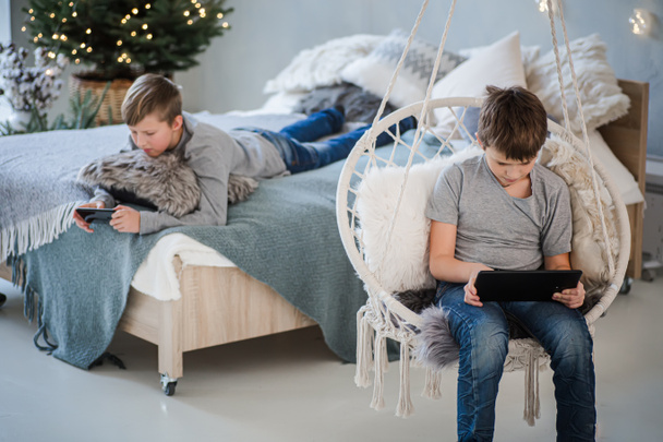 Hermanos charlando a través de gadgets mientras esperan la Navidad cerca del árbol de Navidad en una acogedora casa. - Foto, Imagen