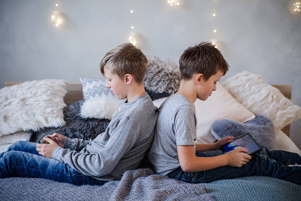 Δύο αγόρια που κάθονται με την πλάτη γυρισμένη ο ένας στον άλλο και παίζουν παιχνίδια στα κινητά τους με φόντο τα χριστουγεννιάτικα φώτα στον τοίχο. - Φωτογραφία, εικόνα