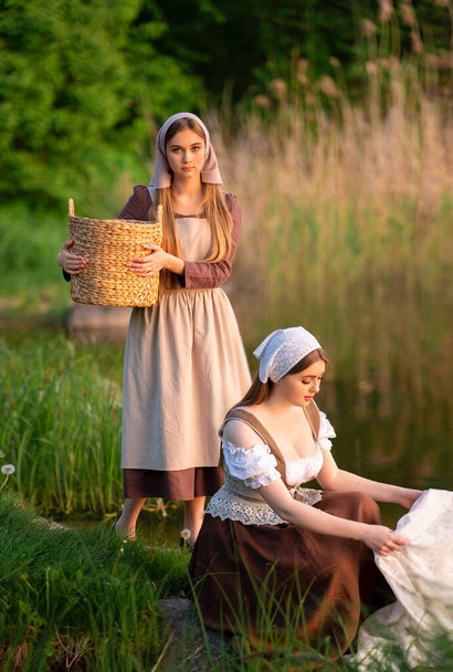 Kaunis nuori kaksi pyykkiä keskiajan puku pesu mekot lähellä jokea. Kaunis tyttö työskentelee maaseudulla. Satutaideteos. - Valokuva, kuva