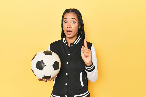Indonesian schoolgirl with soccer ball on yellow having an idea, inspiration concept. - Photo, Image