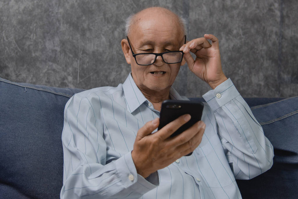 Aziatische oudere man praat mobiele telefoon thuis bank. Technologische communicatie en sociaal netwerkconcept. - Foto, afbeelding