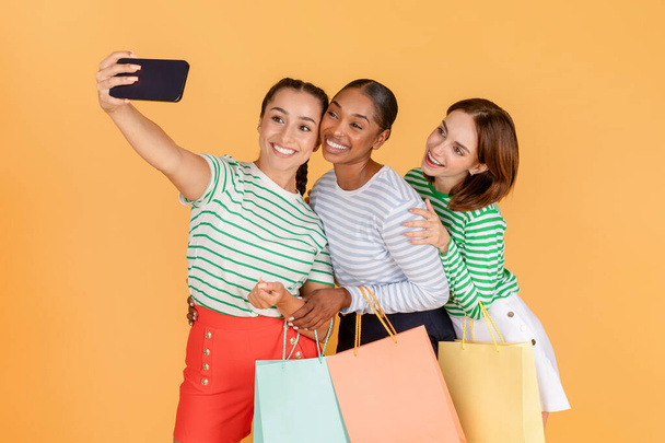 Χαριτωμένη όμορφη νεαρή πολυφυλετικές κυρίες shopaholics λαμβάνοντας selfie μαζί στο κινητό τηλέφωνο απολαμβάνοντας ψώνια, απομονώνονται σε πορτοκαλί φόντο. Αναψυχή, ταξίδια, χαρούμενη έννοια του τρόπου ζωής - Φωτογραφία, εικόνα