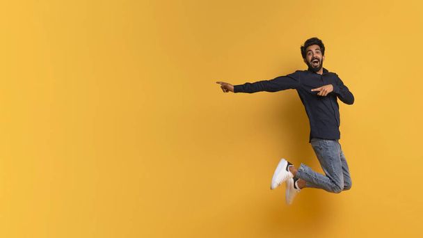 Great Offer. Excited indian man pointing aside while jumping up in air, positive young eastern male having fun and demonstrating free copy space on yellow studio background, panorama - Photo, Image