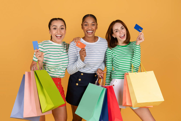 Drie aantrekkelijke jonge multi-etnische dames vriendinnen dragen kleurrijke tassen aankopen, winkelen samen, met behulp van credit bank kaarten, geïsoleerd op oranje achtergrond. Eenvoudig winkelen - Foto, afbeelding