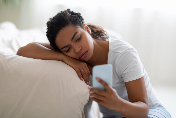 Depressionskonzept. Porträt einer frustrierten afrikanisch-amerikanischen Frau, die aufs Smartphone schaut, aufgebracht soziale Medien checkt, auf dem Boden neben dem Bett sitzt, auf Nachricht oder Anruf wartet, schlechte Nachrichten liest - Foto, Bild
