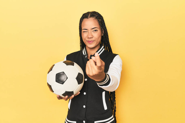 Indonezyjska uczennica z piłką nożną na żółtym wskazując palcem na ciebie, jakby zapraszając zbliżyć. - Zdjęcie, obraz