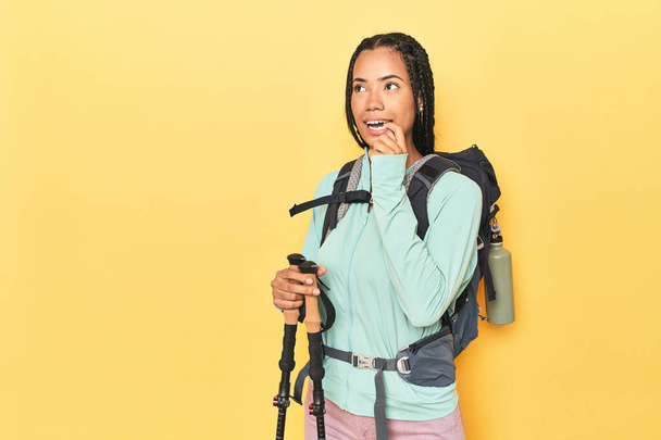 Indonesierin mit Wanderausrüstung auf gelbem Grund denkt entspannt über etwas nach, das einen Kopierraum betrachtet. - Foto, Bild