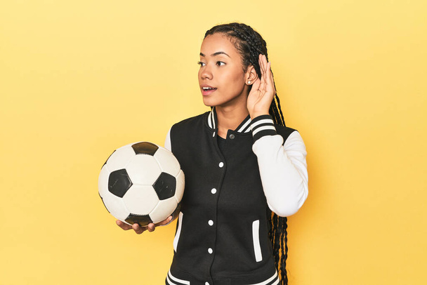 Indonesian schoolgirl with soccer ball on yellow trying to listening a gossip. - Photo, Image